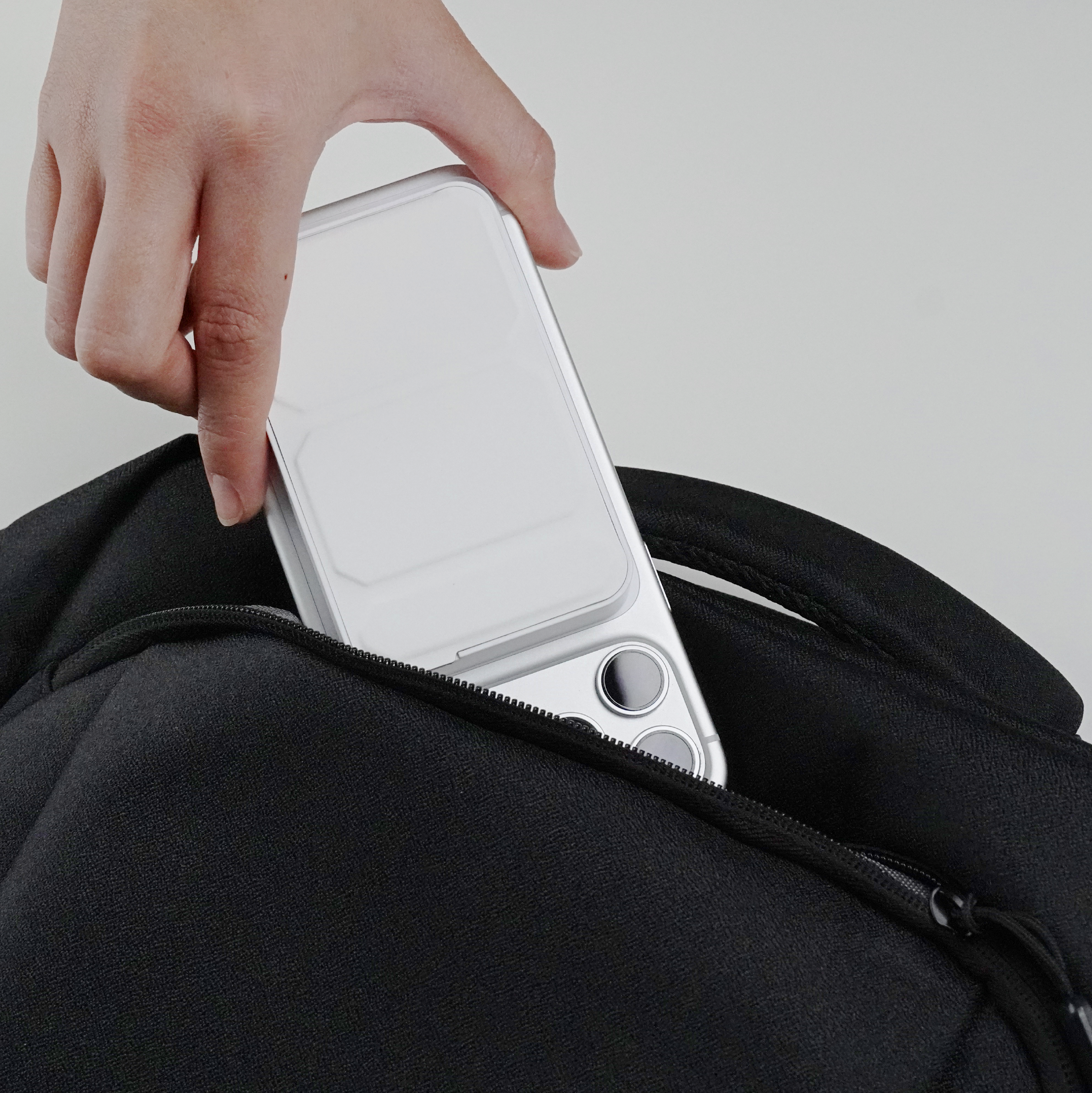 Person placing a smartphone and power bank into a black backpack against a neutral background